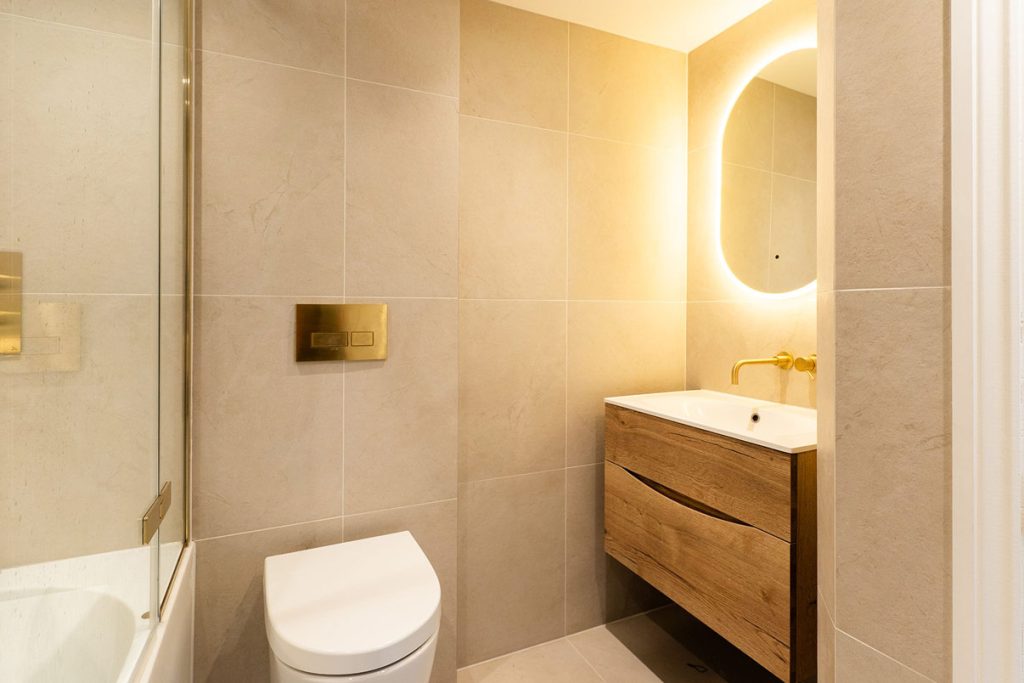 A warm stone-tiled bathroom with a wall-hung oak vanity, a brushed brass wall-mounted tap, a backlit pill-shaped mirror, a brass flush plate and a wall-hung toilet