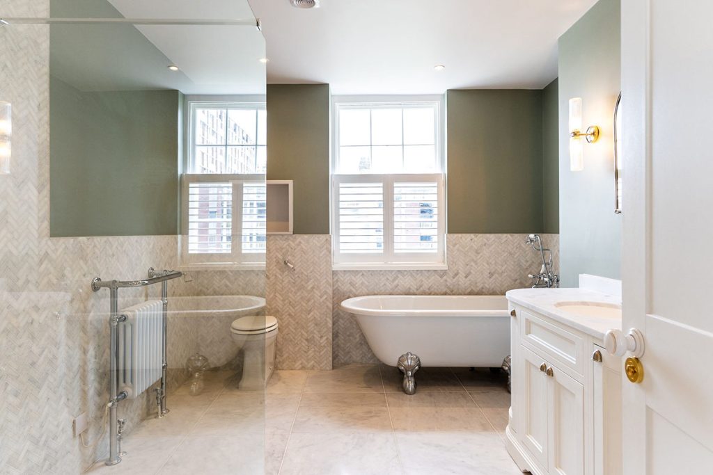 A classic bathroom with a roll-top clawfoot bath, herringbone marble wall tiles below sage green walls, a chrome traditional radiator, a white panelled vanity and shuttered sash windows