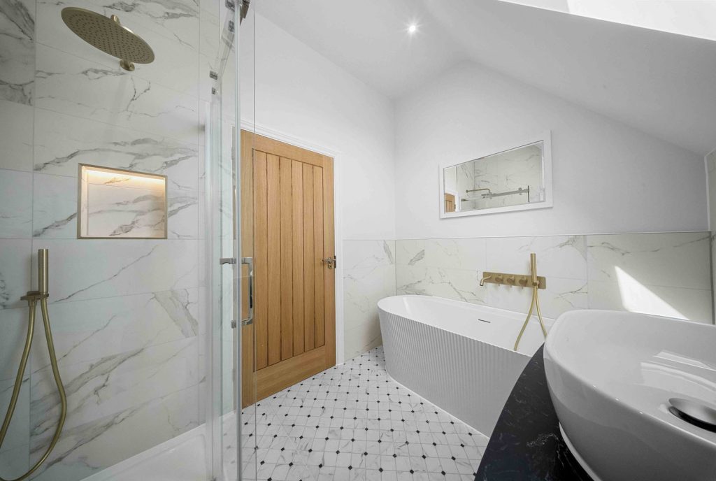 A marble-effect bathroom with a fluted freestanding bath, brushed brass wall-mounted taps, a walk-in shower with a lit niche, an oak plank door and a patterned mosaic floor
