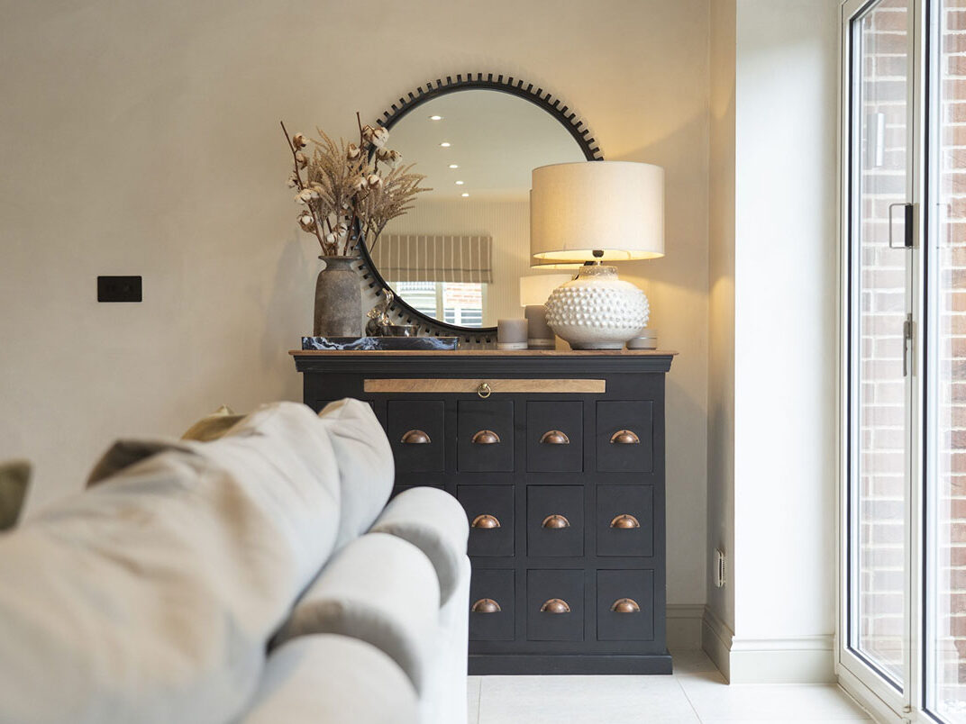 Table lamp casting a warm glow on a dark apothecary chest beside a round mirror, in a Moor House living room by Styled